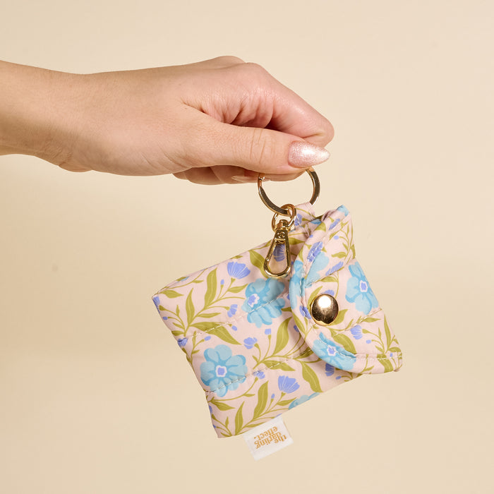 A hand holds The Darling Effect Poppin' Petals Aqua Puffy Earbud Case—a small fabric pouch for wireless earbuds, with blue and green floral print, gold keychain and clasp, and a secure button closure.