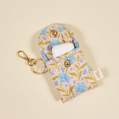 The Darling Effect's Poppin' Petals Aqua Puffy Earbud Case features a blue floral design, gold snap buttons, and a keychain. It securely holds an open white wireless earbuds case inside.