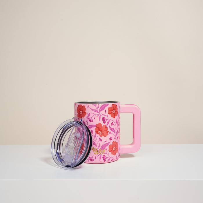 The Darling Effect's Poppin' Petals Hot Pink 14 oz Insulated Mug has double-wall insulation, a large handle, and a floral red/pink pattern. Photographed on a white surface with its clear plastic lid; plain beige background.