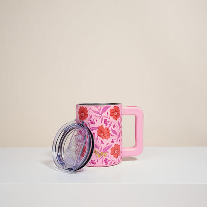The Darling Effect's Poppin' Petals Hot Pink 14 oz Insulated Mug has double-wall insulation, a large handle, and a floral red/pink pattern. Photographed on a white surface with its clear plastic lid; plain beige background.