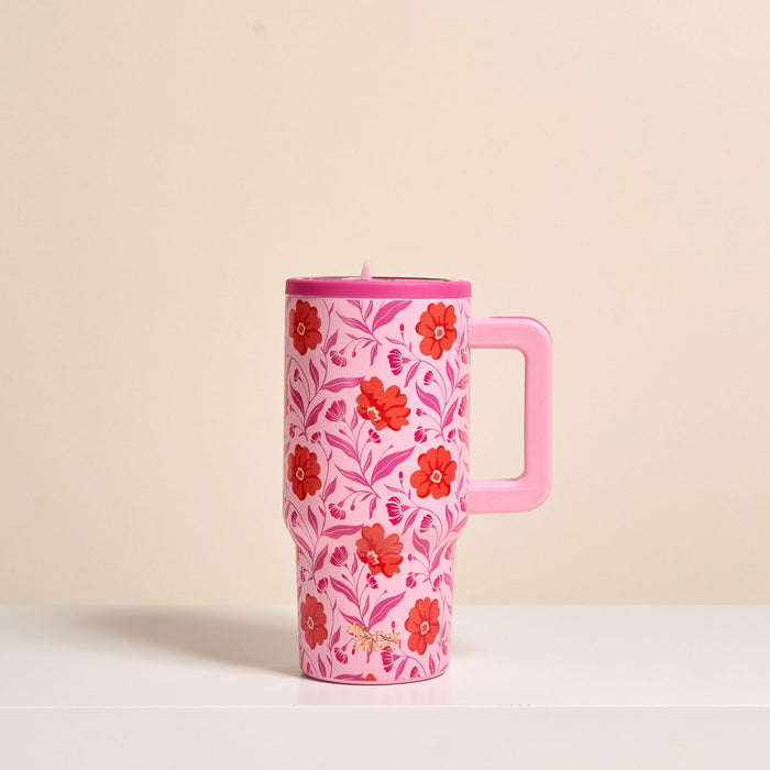 The Darling Effect’s Poppin' Petals Hot Pink 20 oz Lifestyle Flip Straw Tumbler, featuring red and pink flowers with green leaves and a handle, rests on a white surface against a beige background.