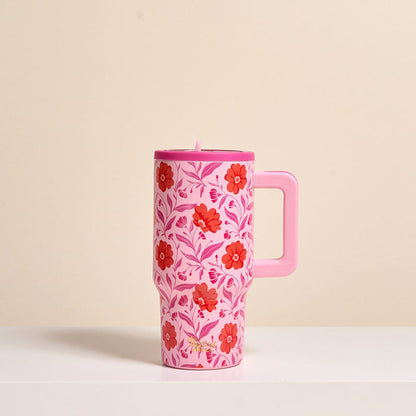 The Darling Effect’s Poppin' Petals Hot Pink 20 oz Lifestyle Flip Straw Tumbler, featuring red and pink flowers with green leaves and a handle, rests on a white surface against a beige background.