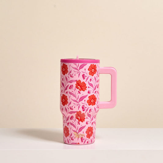 The Darling Effect’s Poppin' Petals Hot Pink 20 oz Lifestyle Flip Straw Tumbler, featuring red and pink flowers with green leaves and a handle, rests on a white surface against a beige background.