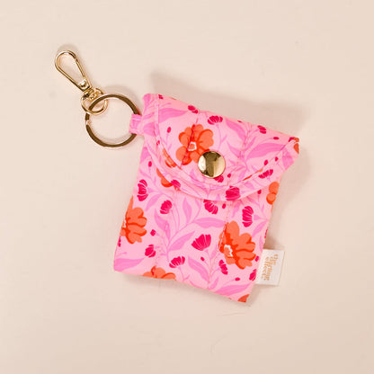 The Darling Effect's Poppin' Petals Hot Pink Puffy Earbud Case features a small, pink floral design, snap button closure, gold keychain attachment, and white fabric tag. Shown on a light background.