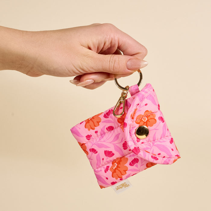 A hand holds The Darling Effect Poppin' Petals Hot Pink Puffy Earbud Case, a quilted floral-patterned pouch keychain with a gold snap button, against a beige background.