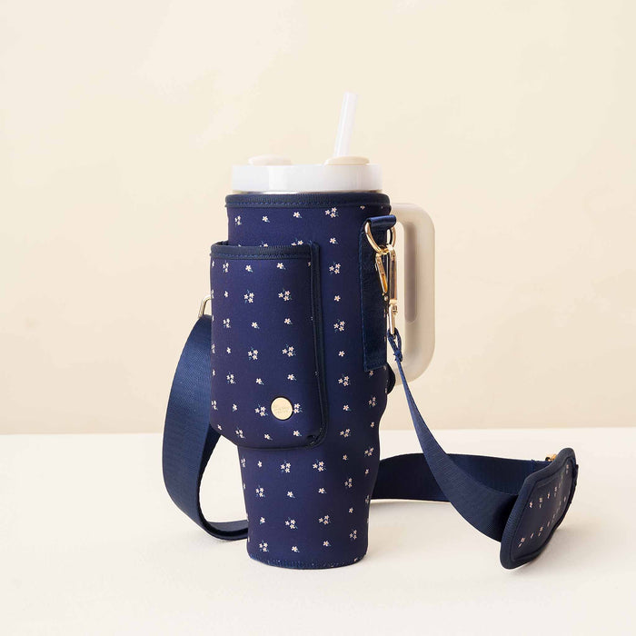 The Darling Effect Pretty Posies Navy Tumbler Sling, a floral-patterned neoprene bag with an adjustable strap and side pocket, holds a large white tumbler with straw and is displayed on a beige surface against a light background.