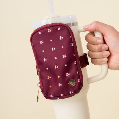 A hand holds a white insulated tumbler with The Darling Effect's Pretty Posies Plum Tumbler Fanny Pack, featuring a water-resistant burgundy material and a small floral pattern.