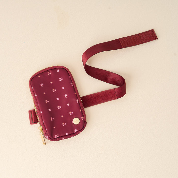 The Darling Effect's Pretty Posies Plum Tumbler Fanny Pack features a maroon pouch with a gold zipper, white floral pattern, and adjustable strap—ideal for carrying daily essentials in water resistant style.