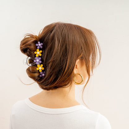 A woman with brown hair in a bun, wearing The Darling Effect's Purple Yellow Game Day Daisy Claw Clip, a white top, and gold hoop earrings, is shown from behind—her look effortlessly nods to school colors against a light background.