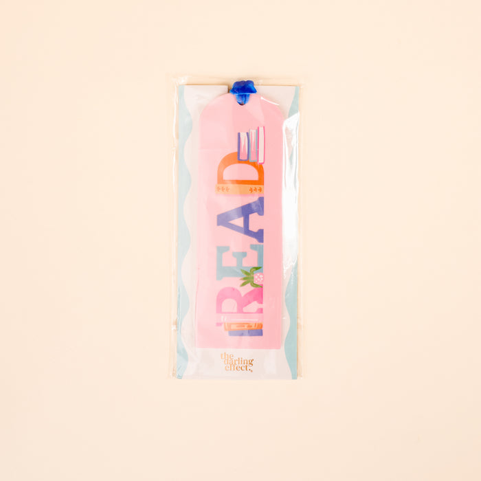 The Darling Effect's Read Acrylic Bookmark features pastel pink acrylic, playful colorful "READ" letters with small illustrations, finished with a velvet ribbon tassel. Comes in a clear plastic sleeve with a cream background.