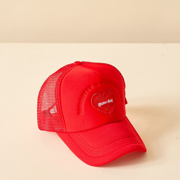 The Darling Effect’s Red Game Day Trucker Hat features a heart-shaped patch with “game day” embroidered in white and a mesh adjustable back. The cap is displayed on a light beige surface with a matching background.
