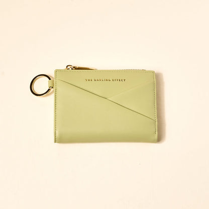 The Sage Luxe Pocket Wallet by The Darling Effect features light green vegan leather, a gold zipper, key ring, and gold-embossed branding—stylish and functional for everyday use, shown here on a beige background.