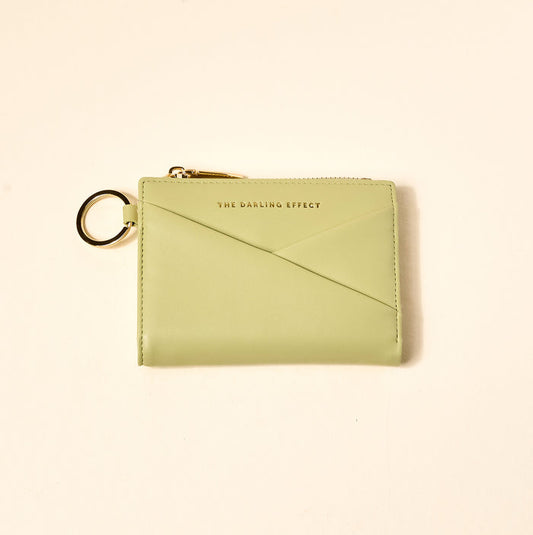 The Sage Luxe Pocket Wallet by The Darling Effect features light green vegan leather, a gold zipper, key ring, and gold-embossed branding—stylish and functional for everyday use, shown here on a beige background.