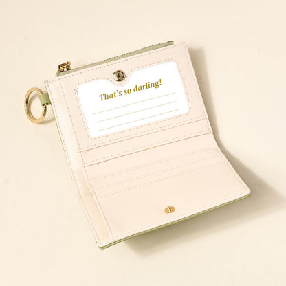 The Darling Effect's Sage Luxe Pocket Wallet is a light pink vegan leather wallet with pale green trim and a keychain. It has a clear ID window with "Thats so darling!", multiple card slots, and is perfect for everyday organization.