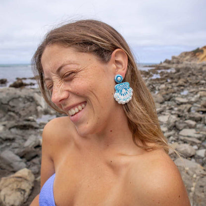A woman laughs with her eyes closed on a rocky beach, wearing The Darling Effect's Seashell Beaded Earrings in playful shapes.