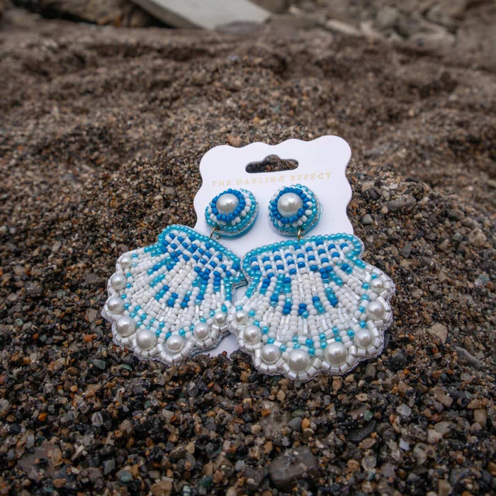 Seashell Beaded Earrings by The Darling Effect showcase vibrant blue and white beads, delicate pearls, and playful seashell shapes, styled on dark sand with tiny pebbles.