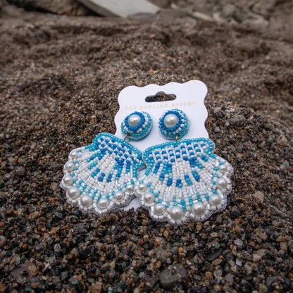 Seashell Beaded Earrings by The Darling Effect showcase vibrant blue and white beads, delicate pearls, and playful seashell shapes, styled on dark sand with tiny pebbles.