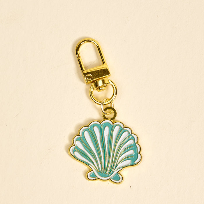 The Darling Effect’s Seashell Pawfect Collar Charm is a gold keychain featuring a blue and white enamel seashell, perfect for adding chic style to your keys or your pet’s collar on a plain light background.