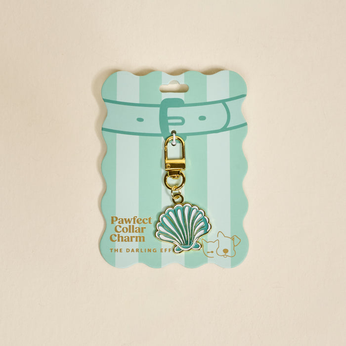 A pastel green, scalloped card from The Darling Effect displays the Seashell Pawfect Collar Charm—a teal and white seashell with a gold clasp. The card features a cat and dog illustration, making it a cute pet accessory addition.
