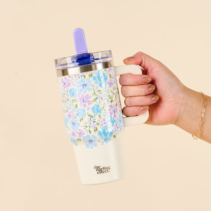A hand holds the-darling-effect Secret Garden Blue 20 oz Lifestyle Flip Straw Tumbler, featuring a blue and purple floral pattern, purple straw, white handle, and leak-resistant design—perfectly stylish and practical for your favorite drinks.