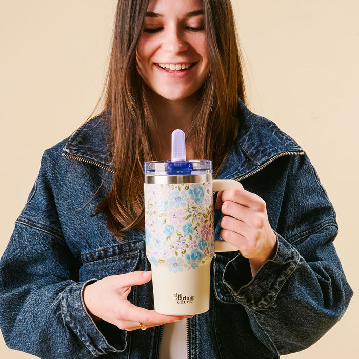 A smiling woman in a denim jacket holds the-darling-effect’s Secret Garden Blue 20 oz Lifestyle Flip Straw Tumbler, featuring a floral pattern, leak-resistant lid, and straw, with "The Thirsty Effect" text against a light beige background.