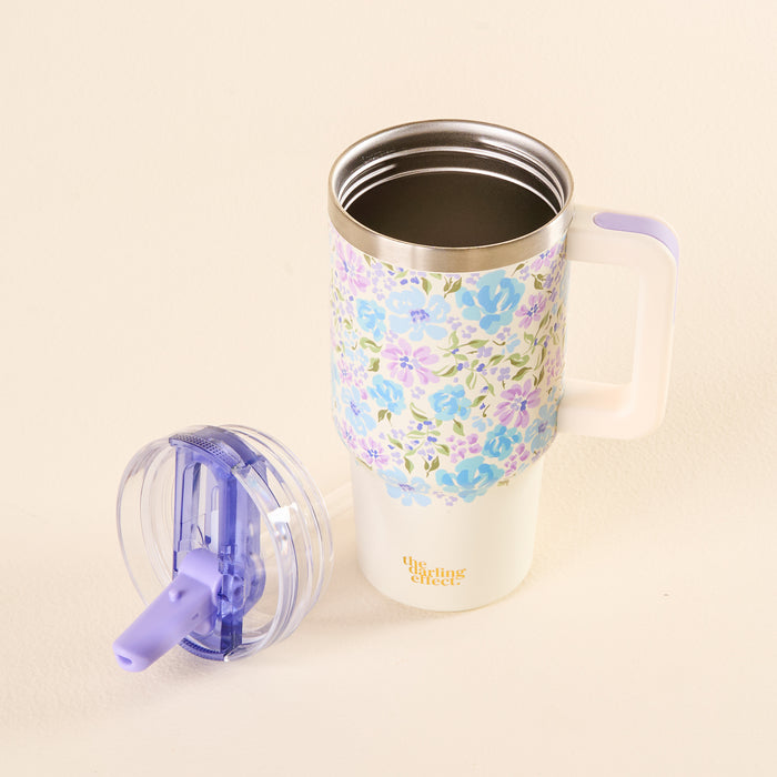 The Secret Garden Blue 20 oz Lifestyle Flip Straw Tumbler from the-darling-effect, with a handle and pastel floral design, sits on a cream surface. Its purple and clear lid is beside it, and “the smiLing effect” is printed in gold on the tumbler.