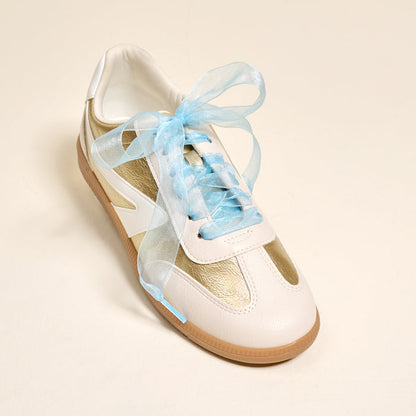 A white and gold sneaker with a gum sole features The Darling Effect’s Sheer Blue Step It Up Shoelaces in place of traditional laces, shown on a plain light background.