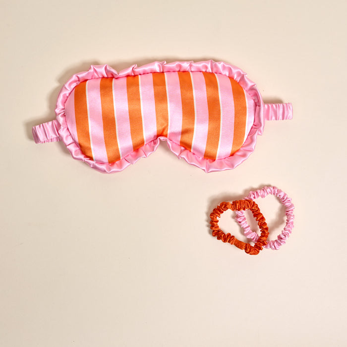 The Sicily Stripes Pink Beauty Sleep Mask by The Darling Effect is paired with pink and orange satin scrunchies, all featuring bold stripes, displayed on a beige background.