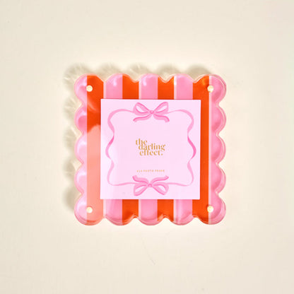 The Sicily Stripes Pink Square Scalloped Acrylic Picture Frame by The Darling Effect features pink and orange stripes, playful scalloped edges, and comes with a decorative brand card, displayed on a light background.