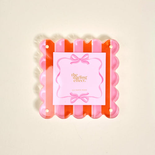 The Sicily Stripes Pink Square Scalloped Acrylic Picture Frame by The Darling Effect features pink and orange stripes, playful scalloped edges, and comes with a decorative brand card, displayed on a light background.