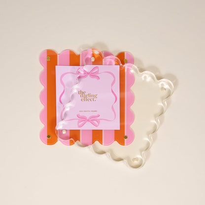 The Darling Effect's Sicily Stripes Pink Square Scalloped Acrylic Picture Frame features pink and orange stripes, gold corner screws, and a clear wavy front for a chic modern look, displayed on a light beige background.