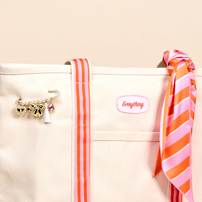 The Darling Effect's Sicily Stripes Pink The Darling Canvas Tote Bag features comfy pink and orange striped straps, a gold safety pin with pearl and charm accents, a pink embroidered Everything patch, and a matching striped scarf tied to the strap.
