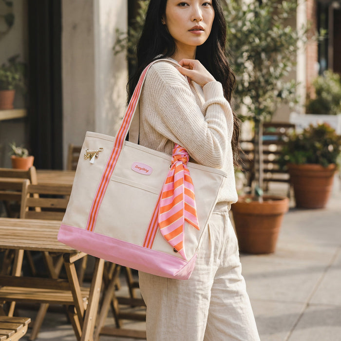 A woman stands outdoors near wooden tables and potted plants, holding The Darling Effect’s Sicily Stripes Pink The Darling Canvas Tote Bag with comfortable straps and a pink striped scarf, paired with light-colored clothes.