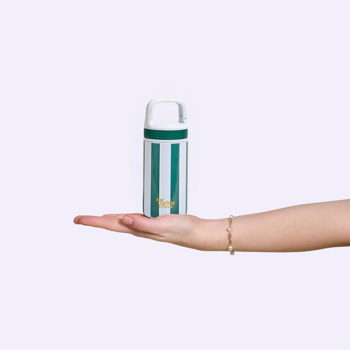 A hand with a bracelet holds The Darling Effect Sicily Stripes Teal 6 oz Lil' Bitty Bottle—a green and white striped water bottle with a white handle—against a plain light background, ideal for hydration on the go.