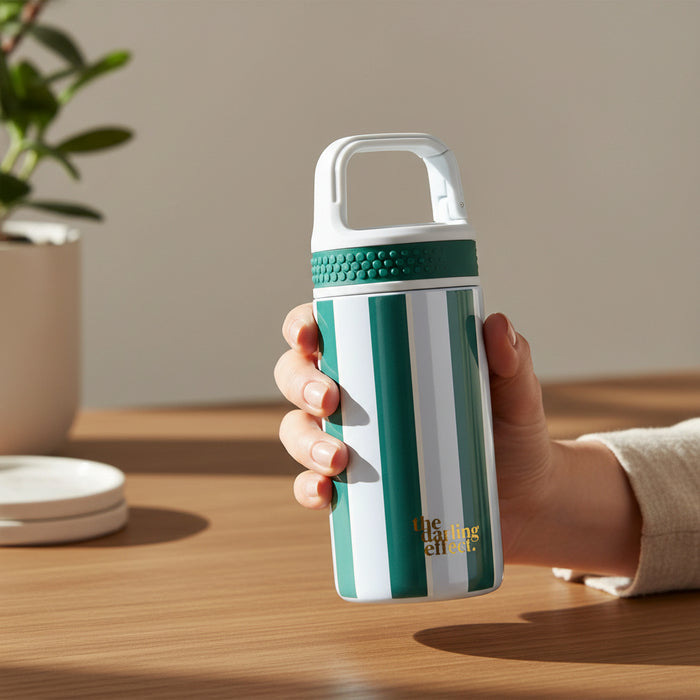 A hand holds The Darling Effect’s Sicily Stripes Teal 6 oz Lil’ Bitty Bottle, featuring a green and white striped design, textured grip, and handle—ideal for on-the-go hydration. A plant and coasters rest on a wooden table nearby.