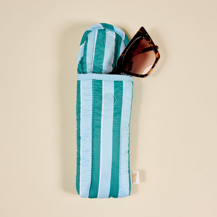 A pair of brown cat-eye sunglasses is partly tucked into The Darling Effect Sicily Stripes Teal Puffy Sunglasses Case, featuring teal and light blue stripes with a magnetic flap closure, set against a beige background.