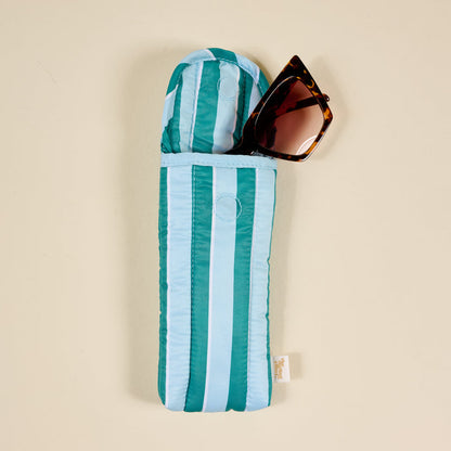 A pair of brown cat-eye sunglasses is partly tucked into The Darling Effect Sicily Stripes Teal Puffy Sunglasses Case, featuring teal and light blue stripes with a magnetic flap closure, set against a beige background.