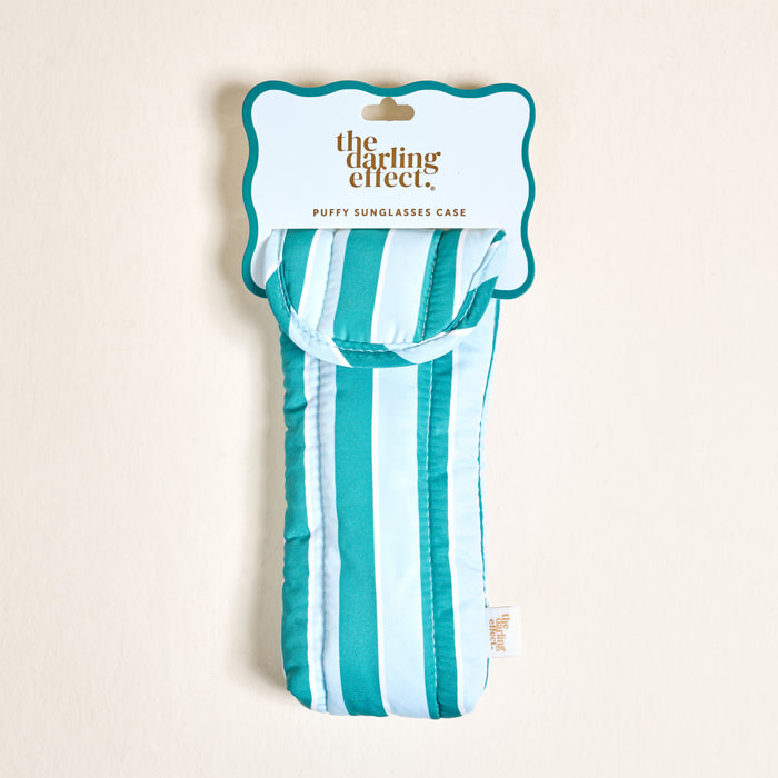The Sicily Stripes Teal Puffy Sunglasses Case by The Darling Effect is shown in its packaging. This stylish case features teal and blue stripes, a magnetic flap closure, and a fabric tag for secure eyeglasses protection.