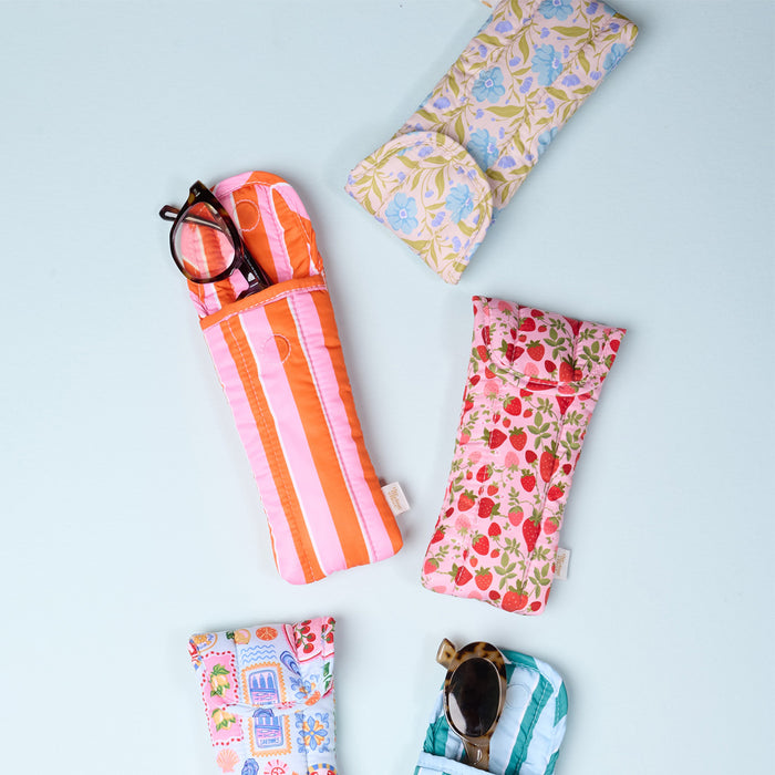 Four soft glasses cases are arranged on a blue background. One Sicily Stripes Teal Puffy Sunglasses Case by The Darling Effect securely holds sunglasses, while another case with a strawberry print provides stylish eyeglasses protection.