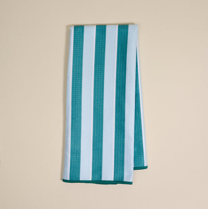 The Darling Effect Sicily Stripes Teal Waffle Tea Towel, featuring wide teal and light blue vertical stripes, is folded and displayed on a plain beige surface.