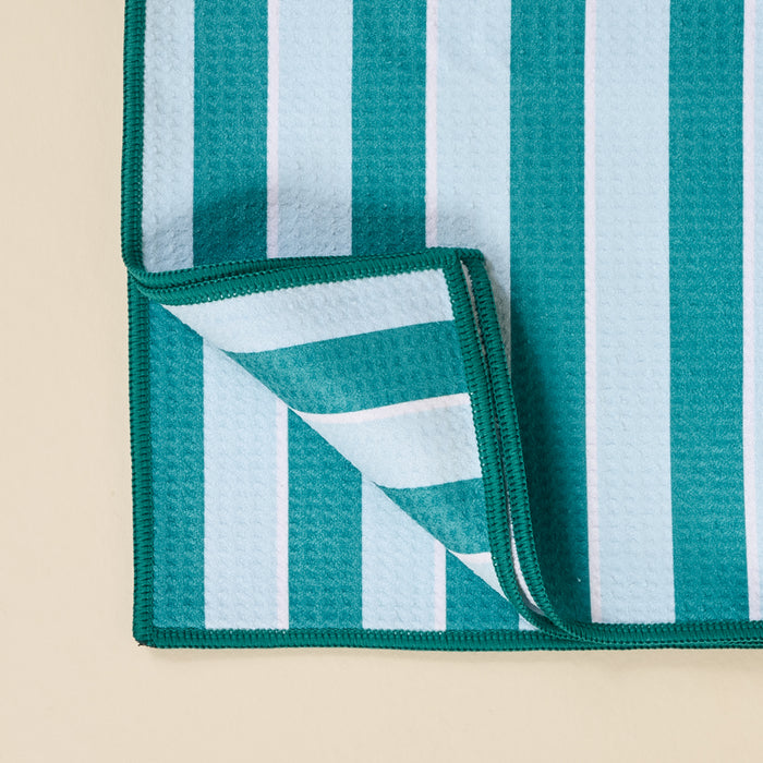 A close-up of The Darling Effect's Sicily Stripes Teal Waffle Tea Towel, featuring absorbent fabric with bold blue, teal, and white stripes, green-edged stitching, and one corner folded to show the underside—an ideal stylish kitchen towel.