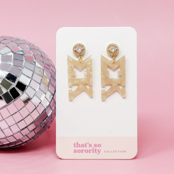 The Darling Effect's Sigma Kappa Letter Gold Acrylic Earrings hang from gold posts with clear gems—perfect for sisterhood events or a bid-day basket. Displayed next to a disco ball on a card reading “thats so sorority COLLECTION.”.