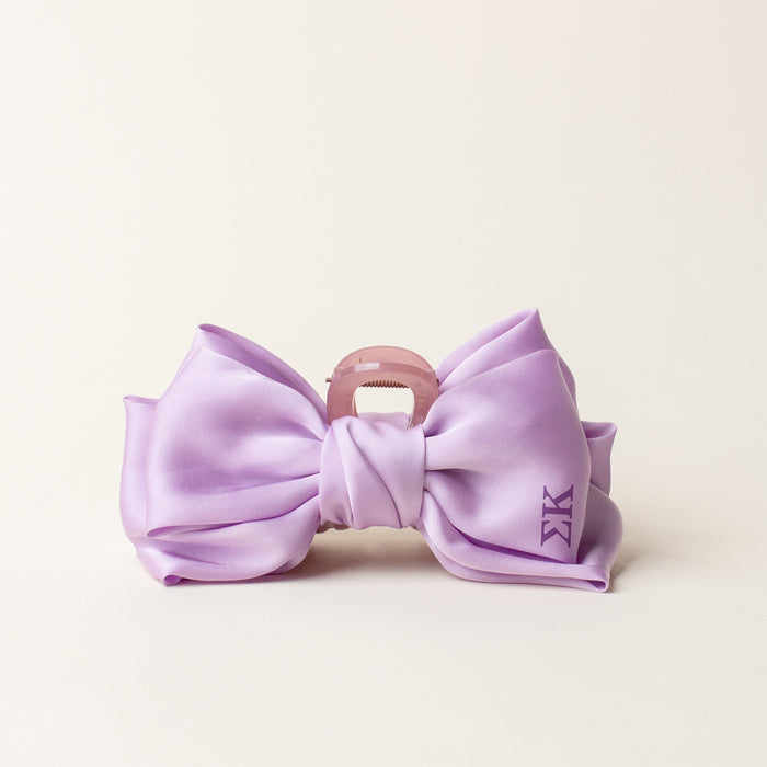 The Darling Effect's Sigma Kappa Satin Bow Claw Clip features a large silky lavender bow with a small KK monogram on the right, attached to a pink claw clip and displayed on an off-white background—perfect for updos.