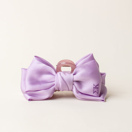 The Darling Effect's Sigma Kappa Satin Bow Claw Clip features a large silky lavender bow with a small KK monogram on the right, attached to a pink claw clip and displayed on an off-white background—perfect for updos.