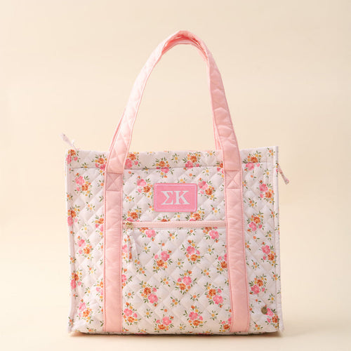 The Darling Effect’s Sigma Kappa The Everyday Quilted Tote Bag features pink handles, floral print, a front pocket, and a pink patch with white ΣΚ letters on beige—perfect for daily use.