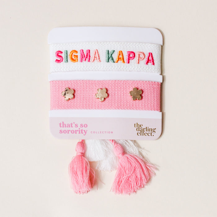 The Sigma Kappa Woven Bracelet Set by The Darling Effect includes a white bracelet with multicolored SIGMA KAPPA letters and a pink bracelet with gold flower studs, both with pink/white tassels. Card reads "that's so sorority" and "The Darling Effect.