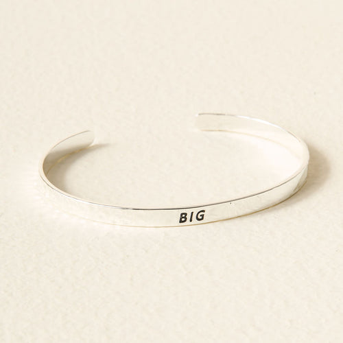 The Darling Effect’s Silver Big Sorority Cuff features “BIG” engraved in black on a simple silver band, shown on a light, textured surface.