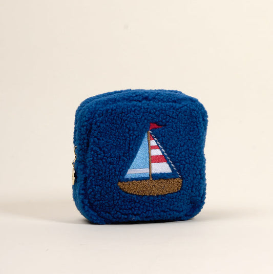The Darling Effect’s Square Sail Boat Teddy Pouch is a small, square blue sherpa zip pouch featuring an embroidered sailboat with colorful sails and a brown hull, shown on a plain off-white background.