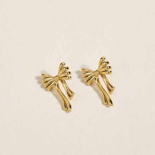 The Darling Effect’s Stay Classy Bow Standout Earrings feature golden, bow-shaped designs with flowing ribbon ends and a high-shine finish, shown on a light beige background.