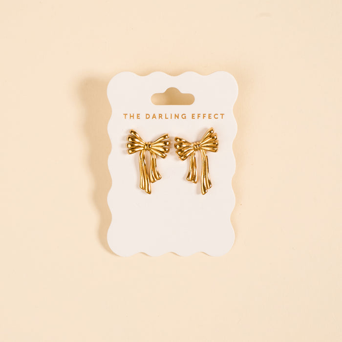 The Darling Effect Stay Classy Bow Standout Earrings, featuring a high-shine gold bow design, are displayed on a branded white card against a beige background.